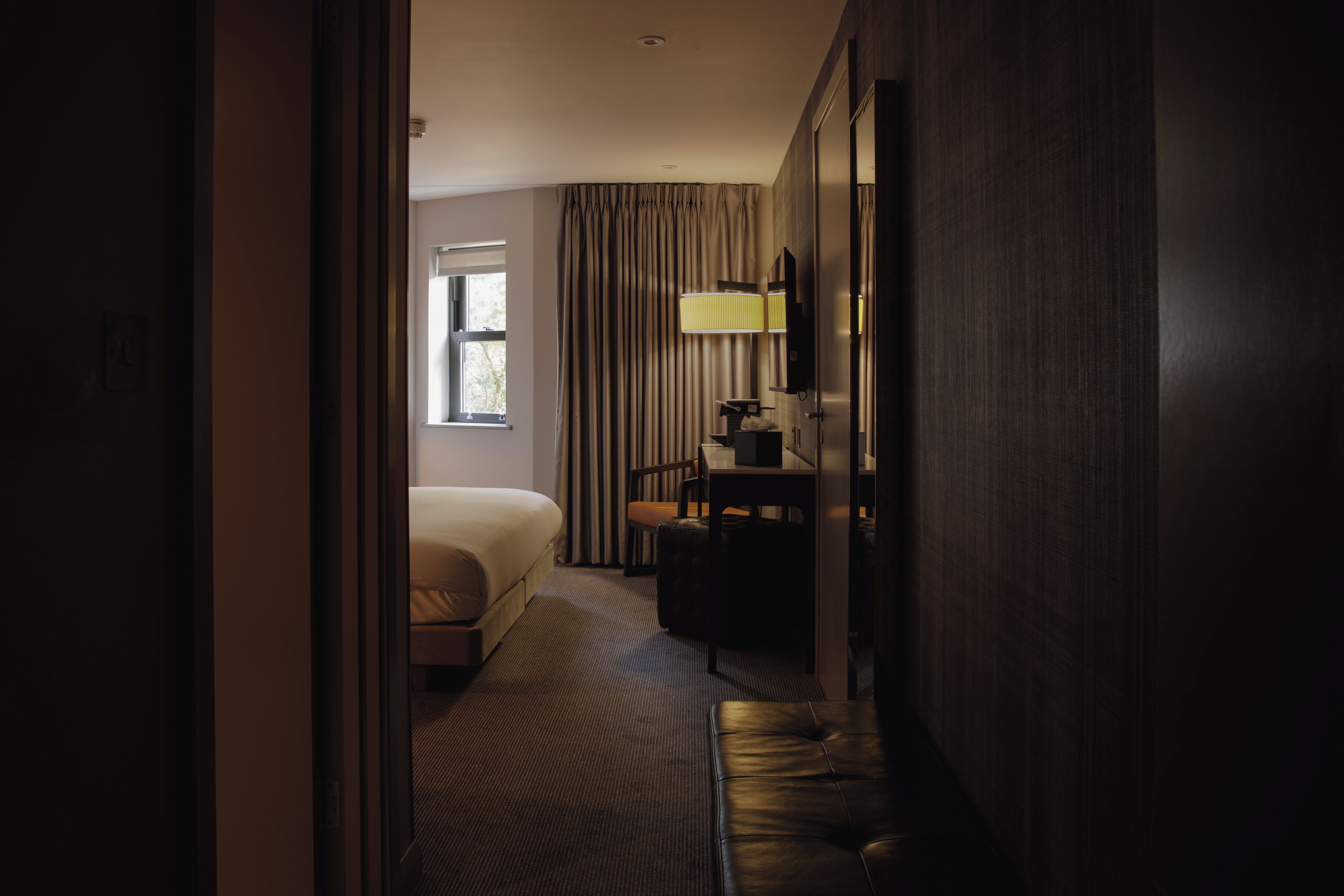 A hotel room with a bed, a desk, a chair, a lamp, and a window.