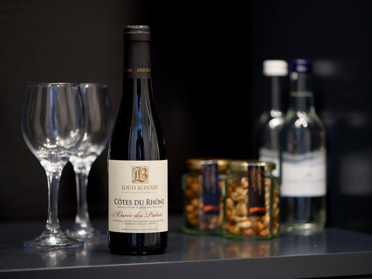 Uma garrafa de vinho Louis Bernard Côtes du Rhône, dois copos de vinho vazios e um pote de nozes sobre uma mesa.