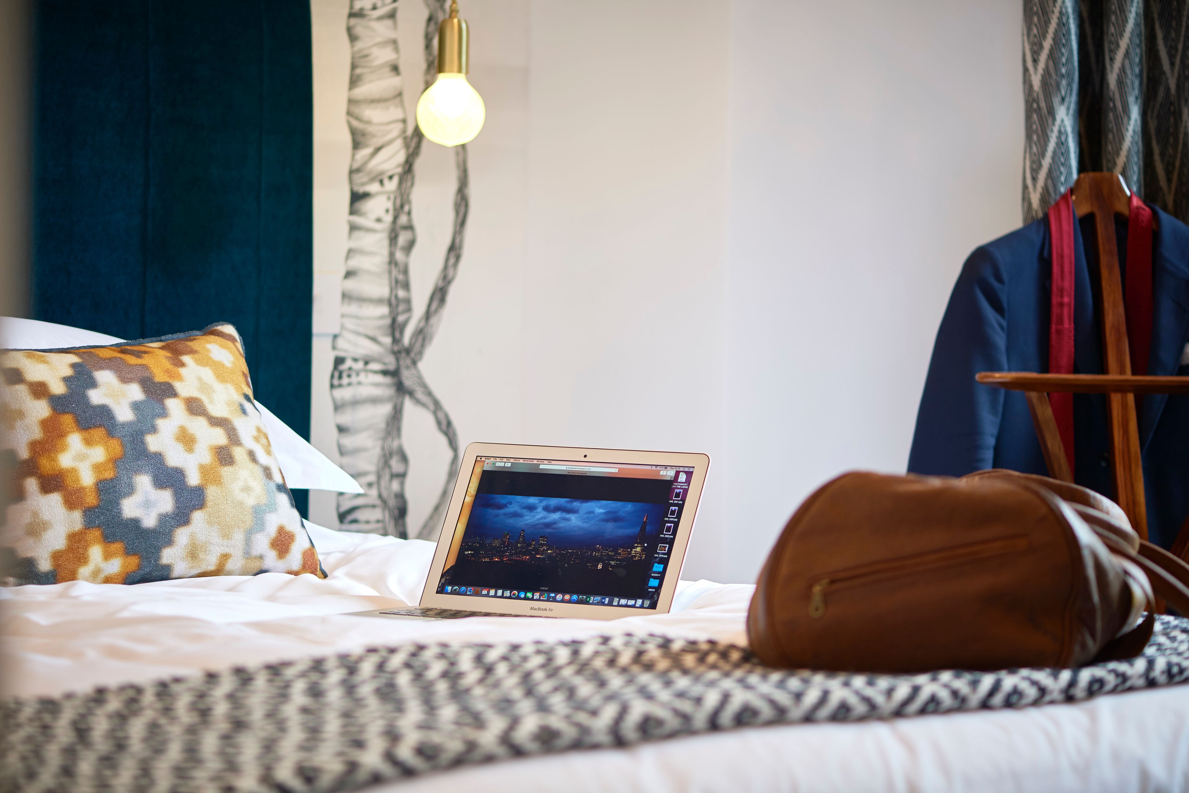A laptop on a bed with a patterned pillow, a brown bag, and a cityscape on the screen.