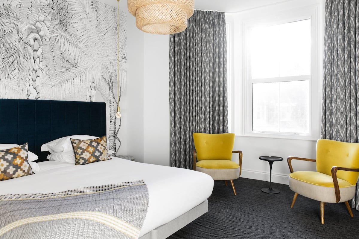 A hotel room with a large bed, two yellow armchairs, a small round table, and a window with curtains.