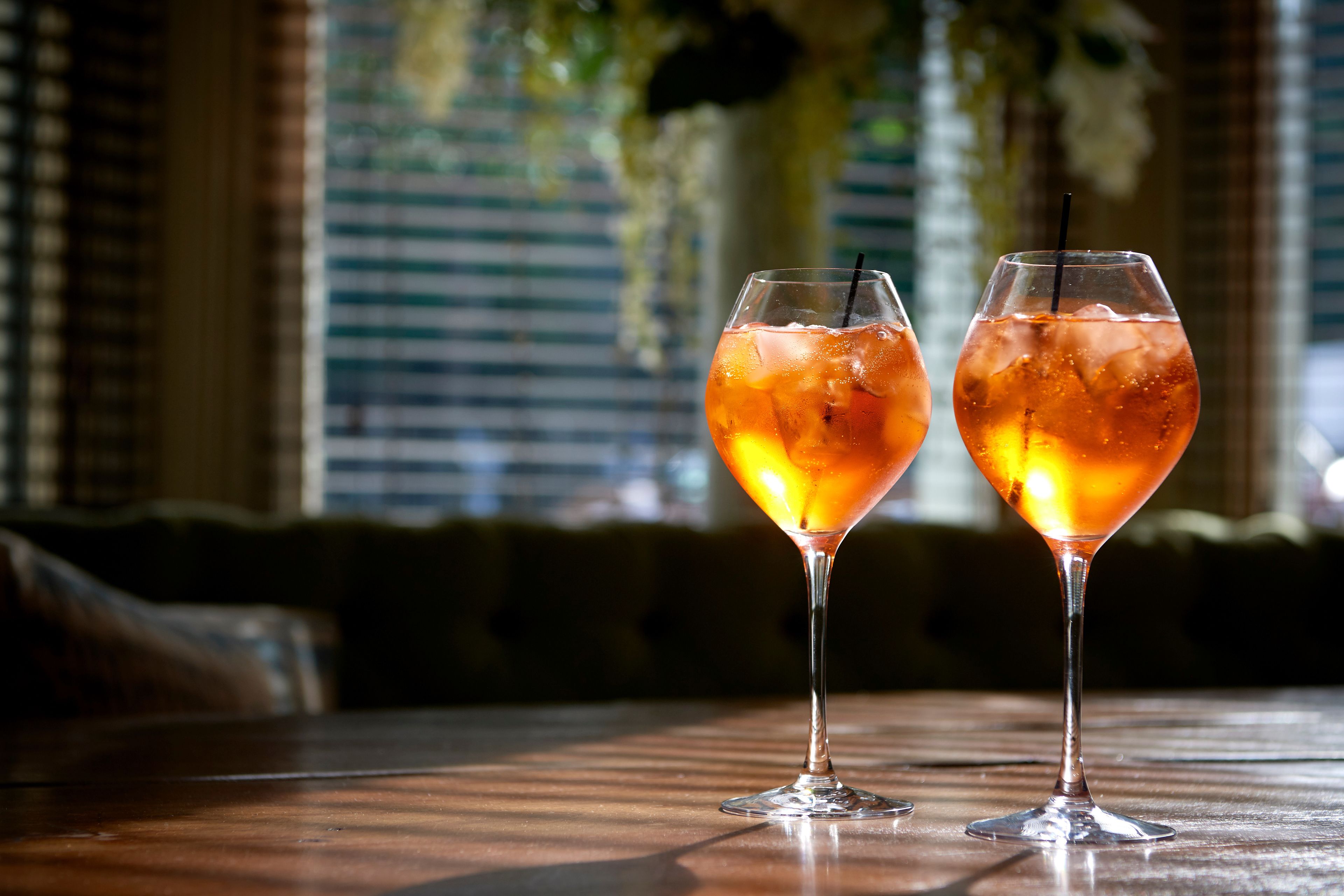 Two glasses of iced tea on a wooden table with a blurred background of a room with a couch and a window with blinds.