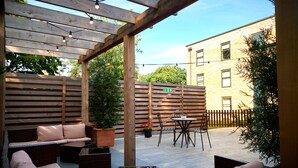 Un patio al aire libre con mobiliario de madera, una pérgola y un edificio de ladrillo al fondo.