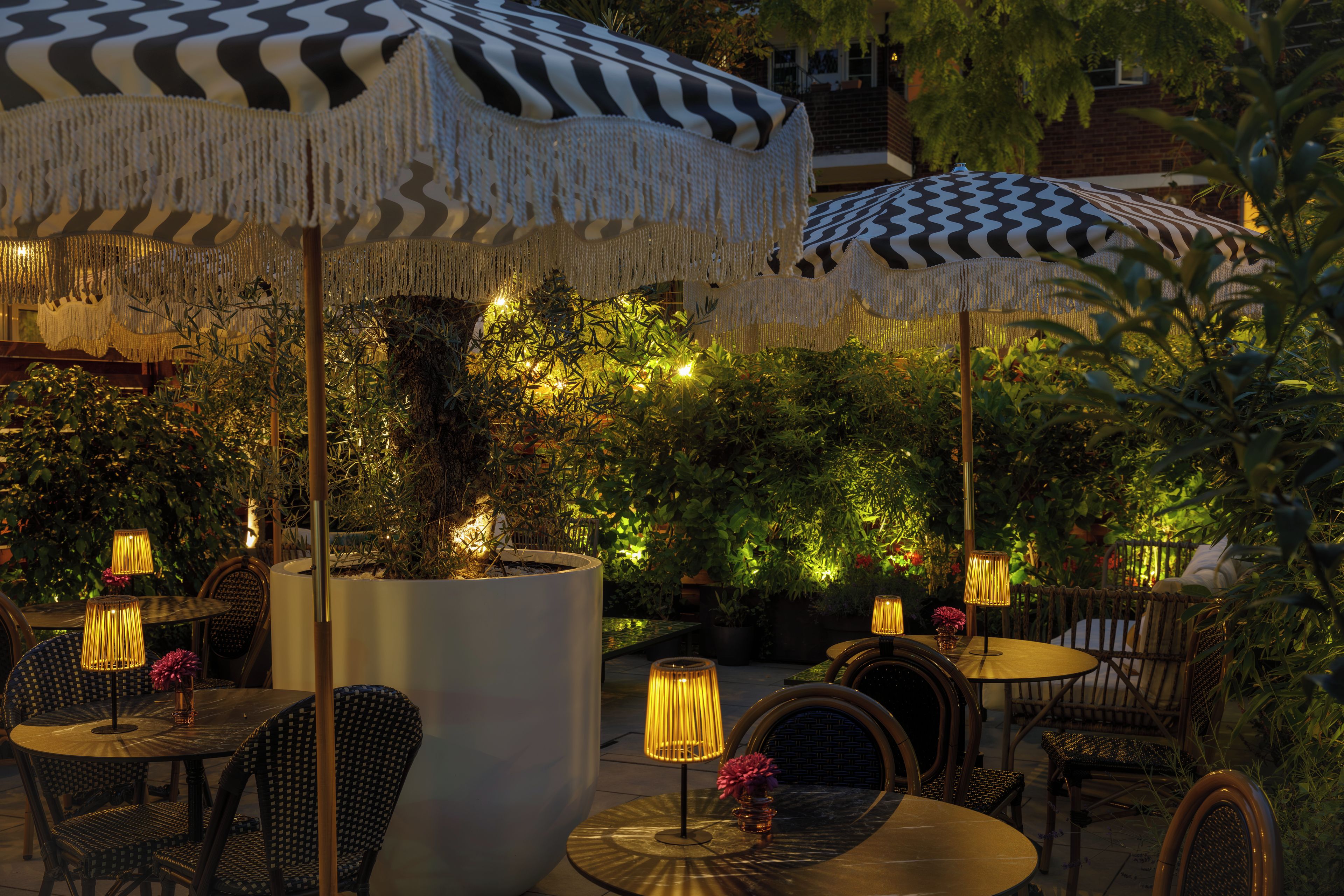 Un espace extérieur aménagé avec des tables et des chaises rondes, protégé par des parasols à motifs et entouré de verdure.
