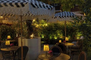 An outdoor seating area with round tables and chairs, covered by patterned umbrellas, and surrounded by greenery. - The Lodge Hotel - Putney (London)