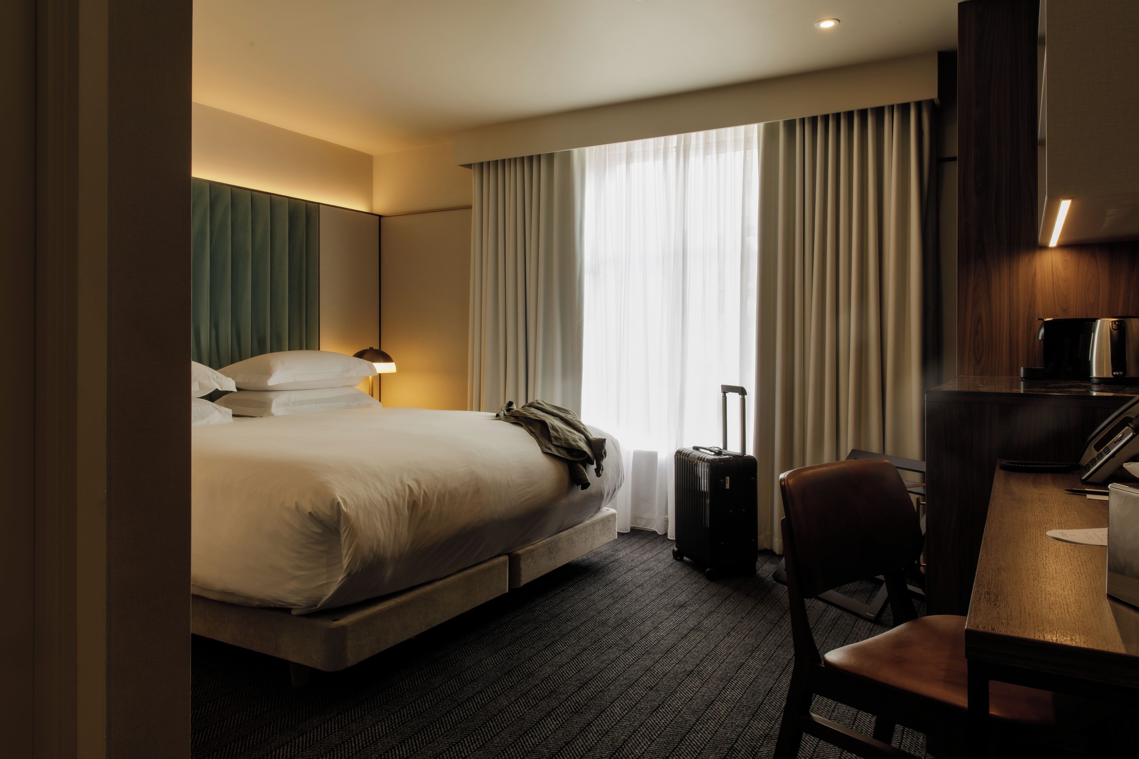 A hotel room with a bed, a desk, a chair, a suitcase, and a coffee machine.