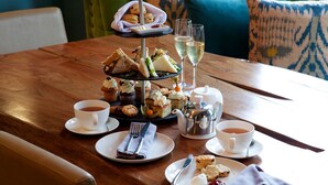 Un expositor de tres niveles con sándwiches, scones y pasteles sobre una mesa de madera, acompañado de tazas de té y copas de vino blanco.