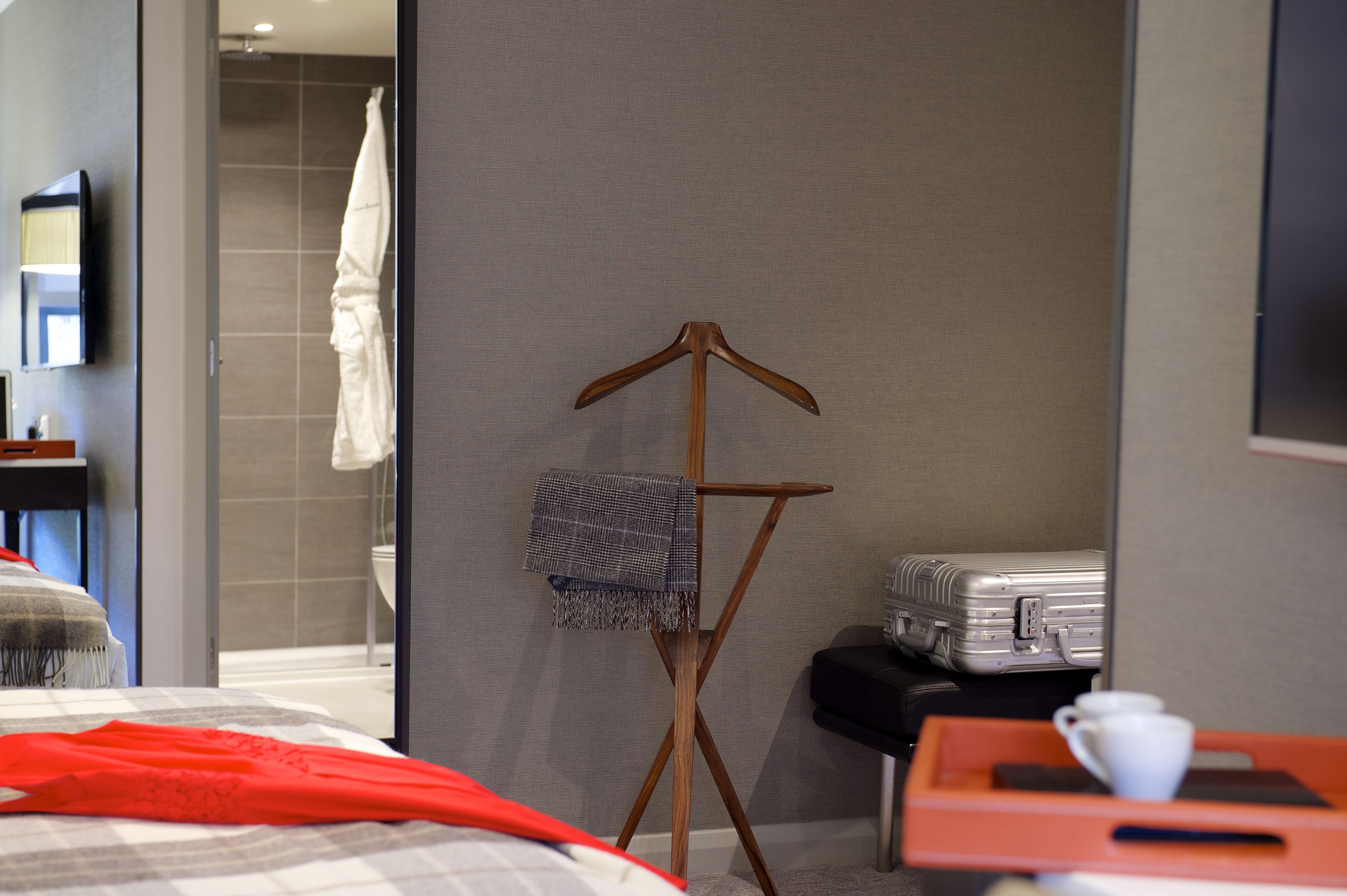 A hotel room with a bed, a wooden folding chair with a blanket, a suitcase, and a tray with a cup.