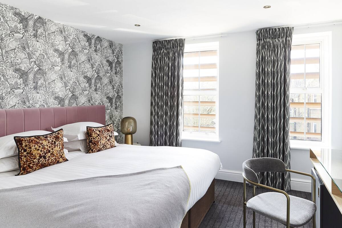 A hotel room with a large bed, a desk, and a chair. The room has a patterned wallpaper, curtains, and a window with a view.