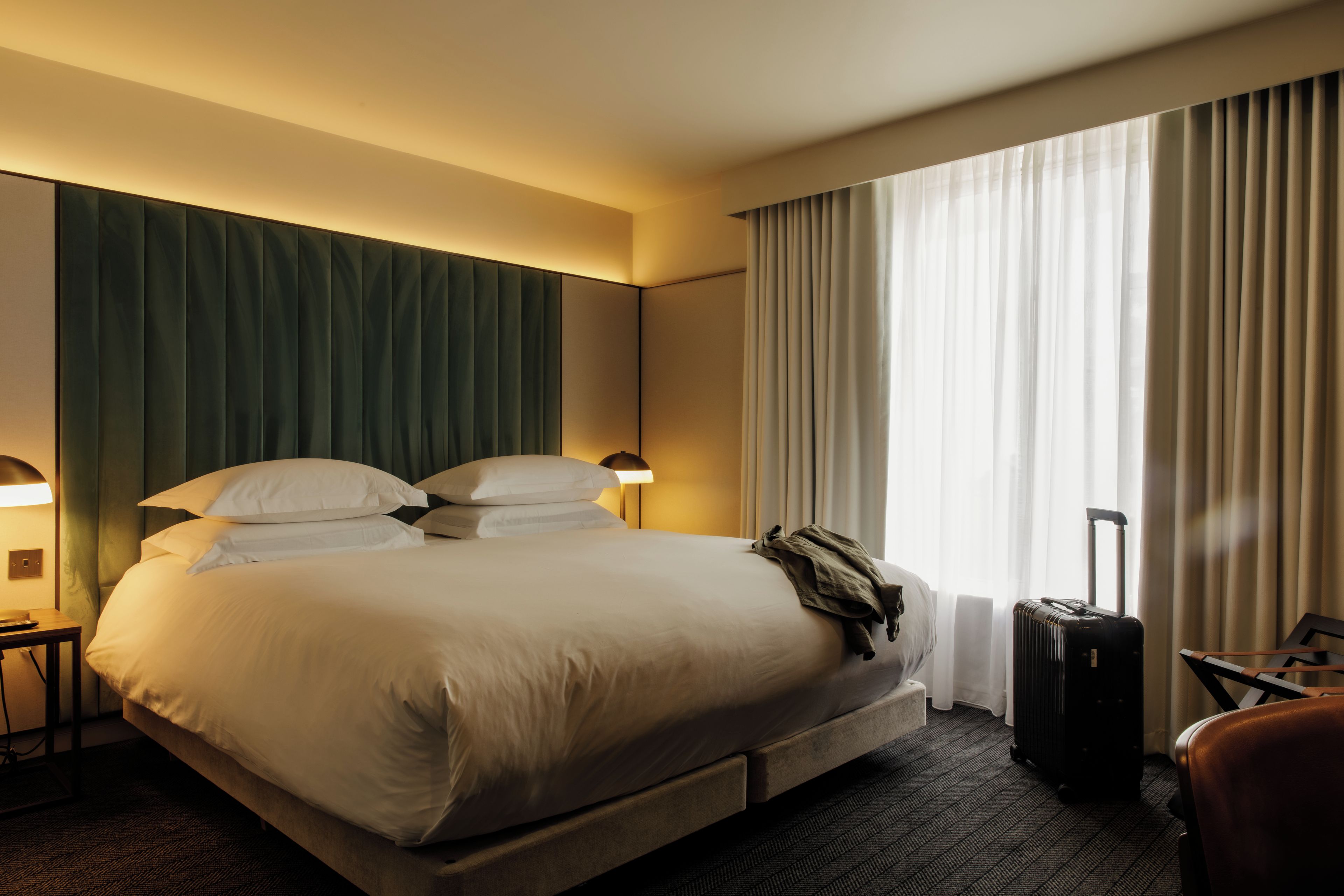 Quarto de hotel com cama grande, mesa de cabeceira, luminária, mala e janela com cortinas.