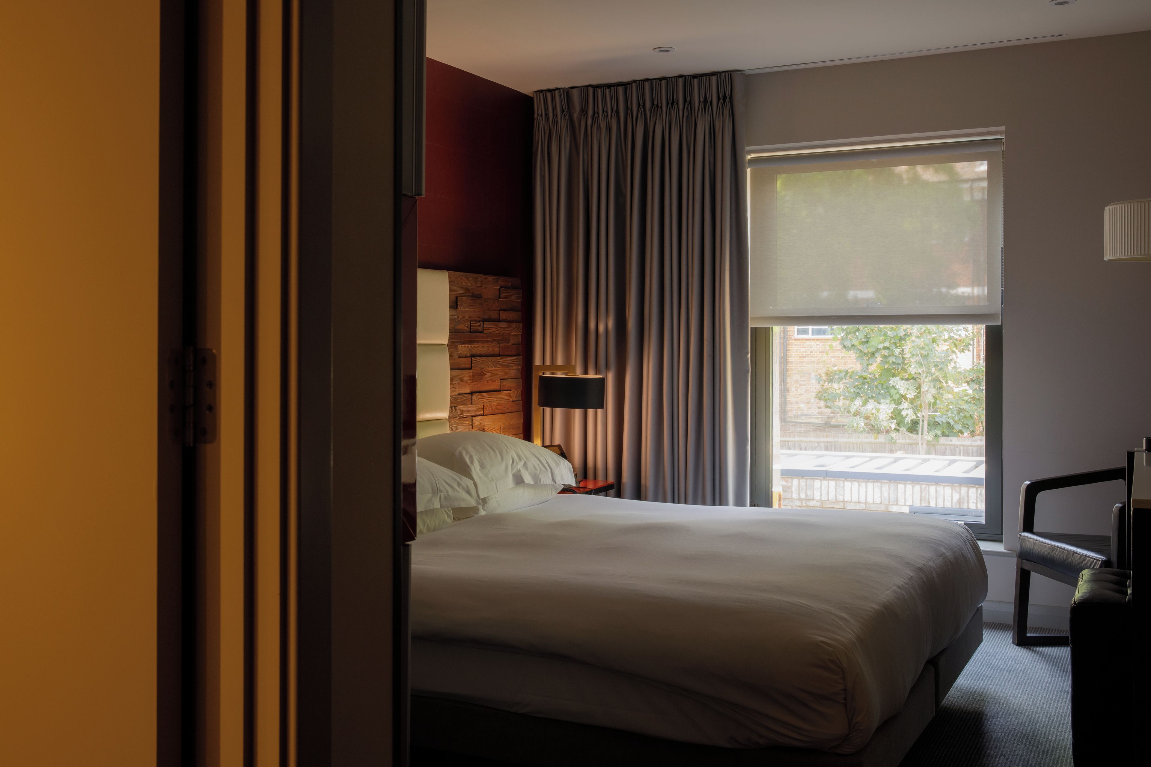 Een hotelkamer met een groot bed, een nachtkastje, een lamp, een raam met gordijnen en uitzicht op buiten.