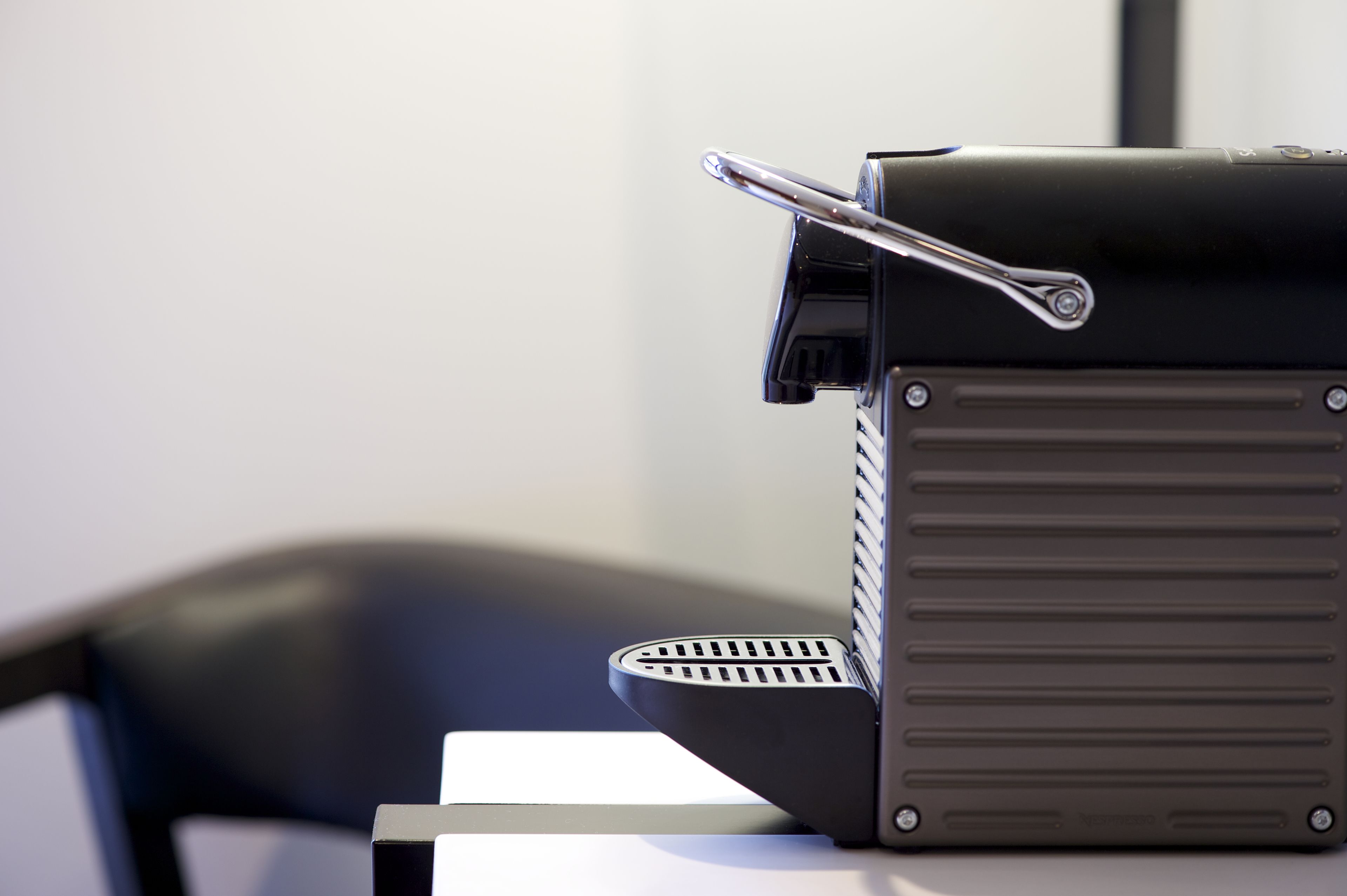 A black portable heater with a metallic handle and vent grilles, placed on a white surface next to a black chair.