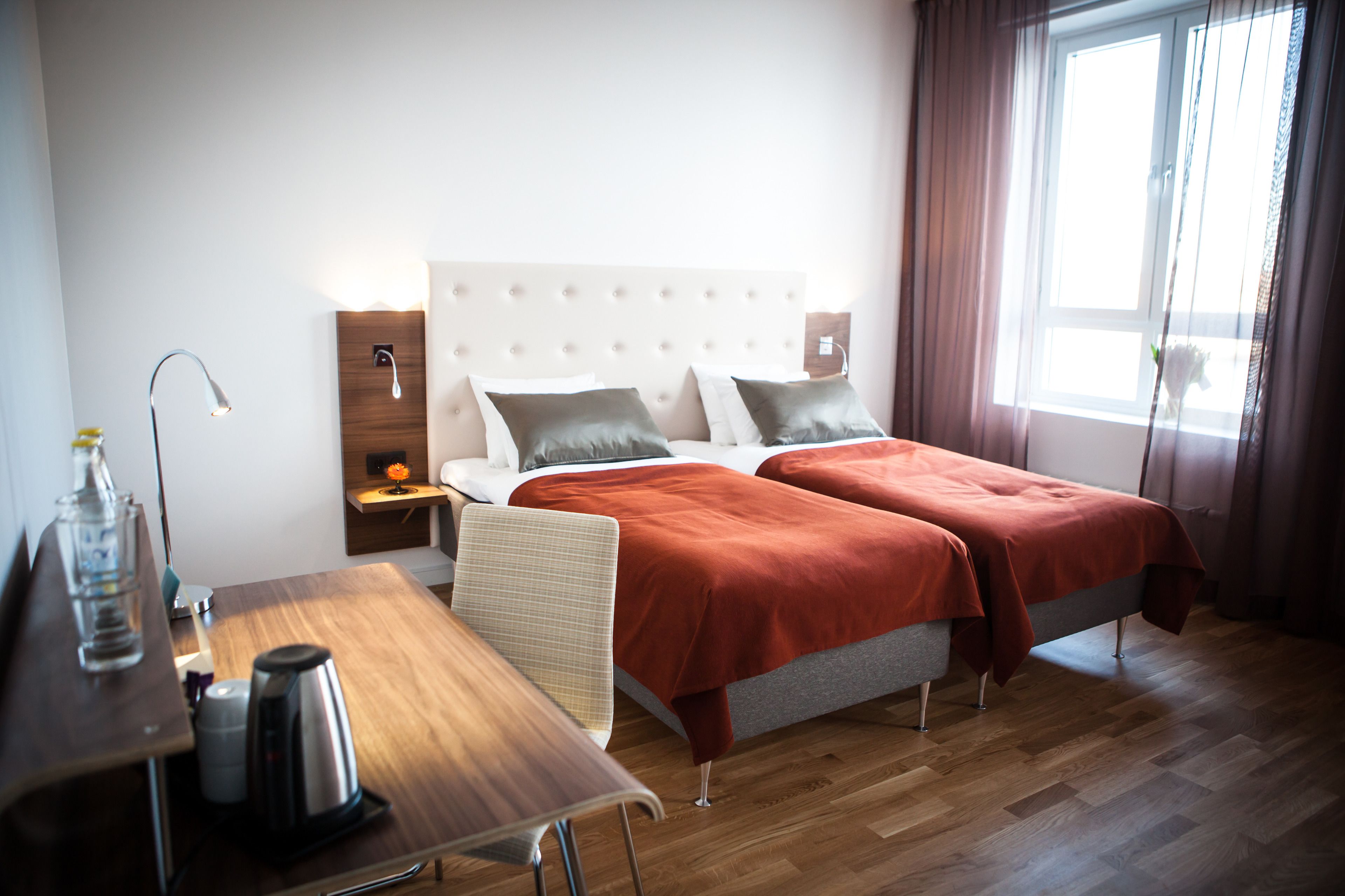 Habitación de hotel con cama doble, mesita de noche con lámpara, una pequeña mesa con hervidor y ventana con cortinas.