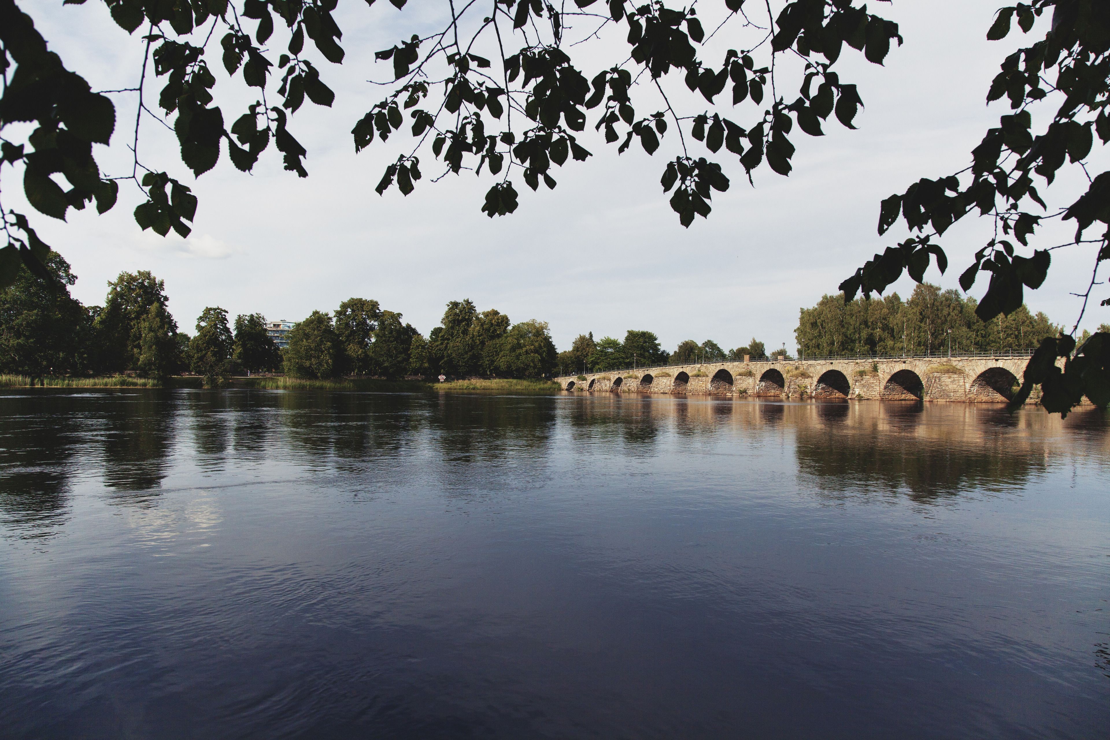 A stone bridge over a calm river with trees in the foreground.
