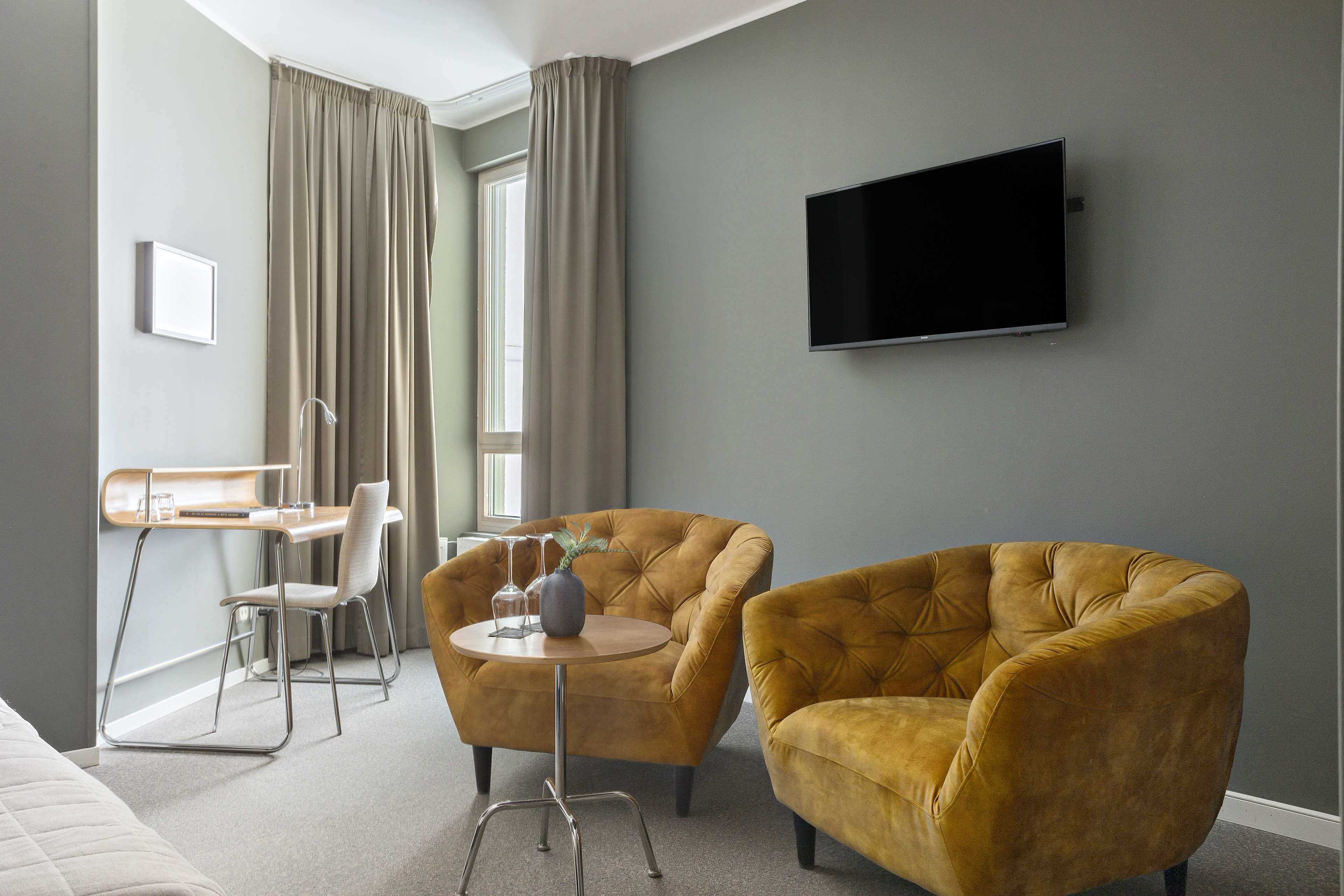 A modern hotel room with a bed, two armchairs, a small round table, a desk with a chair, a flat-screen TV, and a window with curtains.