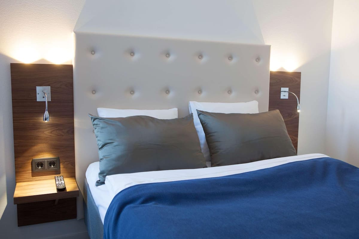 Quarto de hotel com uma cama, uma mesinha de cabeceira com controle remoto e uma luminária fixada na parede.