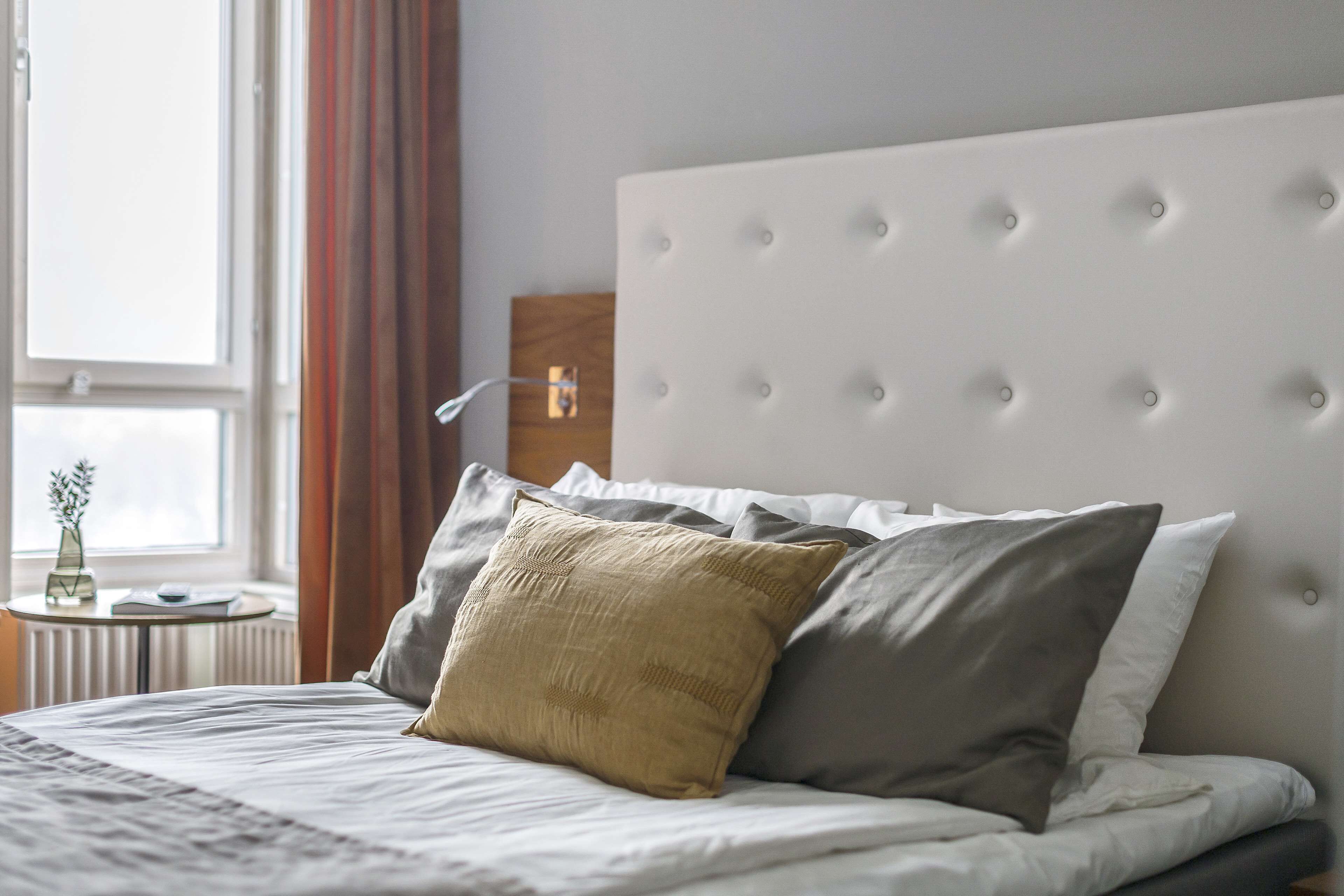 A neatly made bed with a tufted headboard, a bedside table with a vase and book, and a window with curtains.