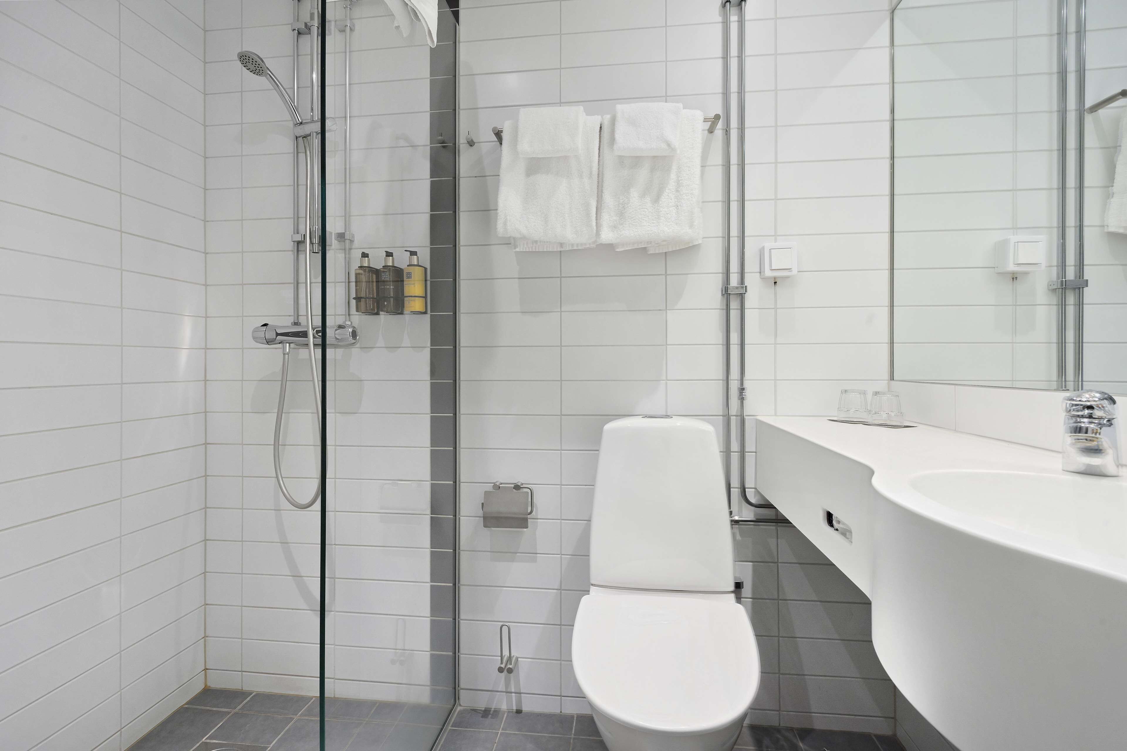 Ett modernt badrum med en dusch som är avskärmad med glasväggar, en vit toalett och ett stort handfat med en spegel.