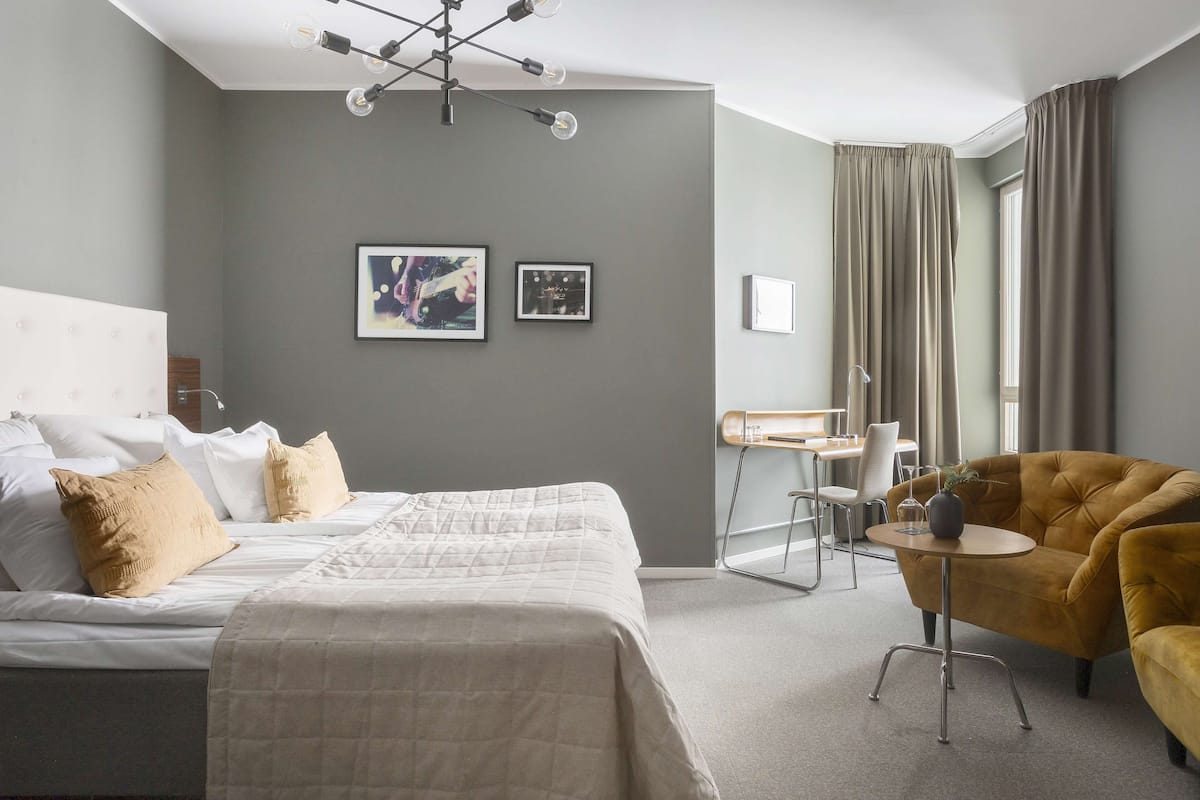 Quarto de hotel moderno com uma cama grande, um sofá, uma mesinha e uma escrivaninha. Há duas imagens emolduradas na parede e uma janela com cortinas.