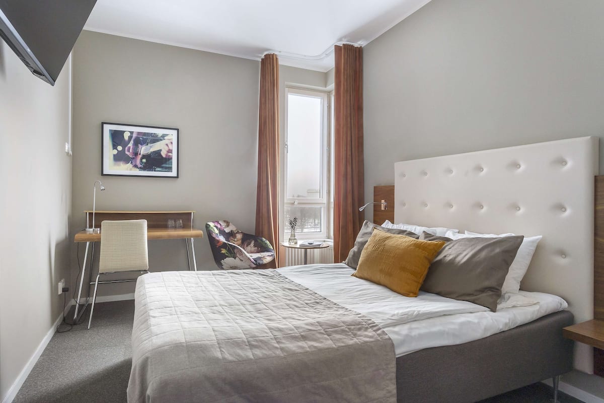 Quarto de hotel com cama, escrivaninha, cadeira e janela com cortinas.