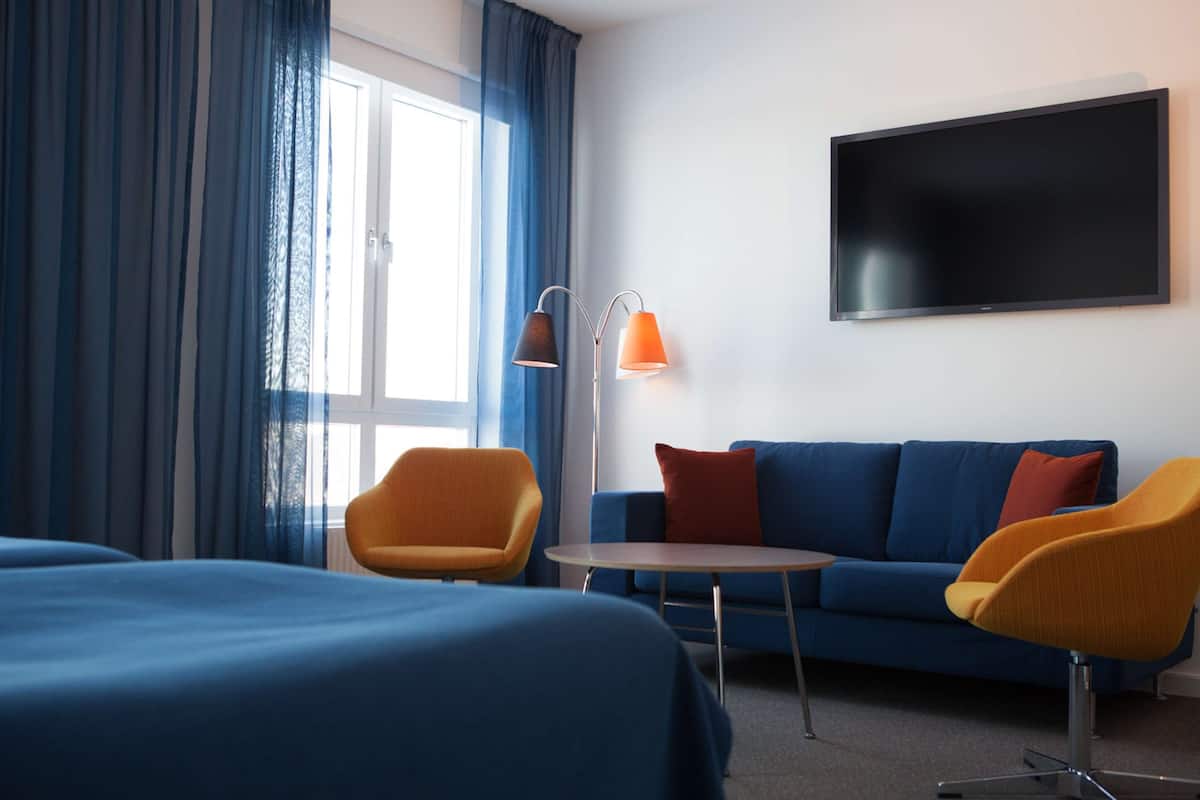 Quarto de hotel com uma cama, um sofá azul, duas poltronas, uma mesa pequena e uma televisão de tela plana na parede.