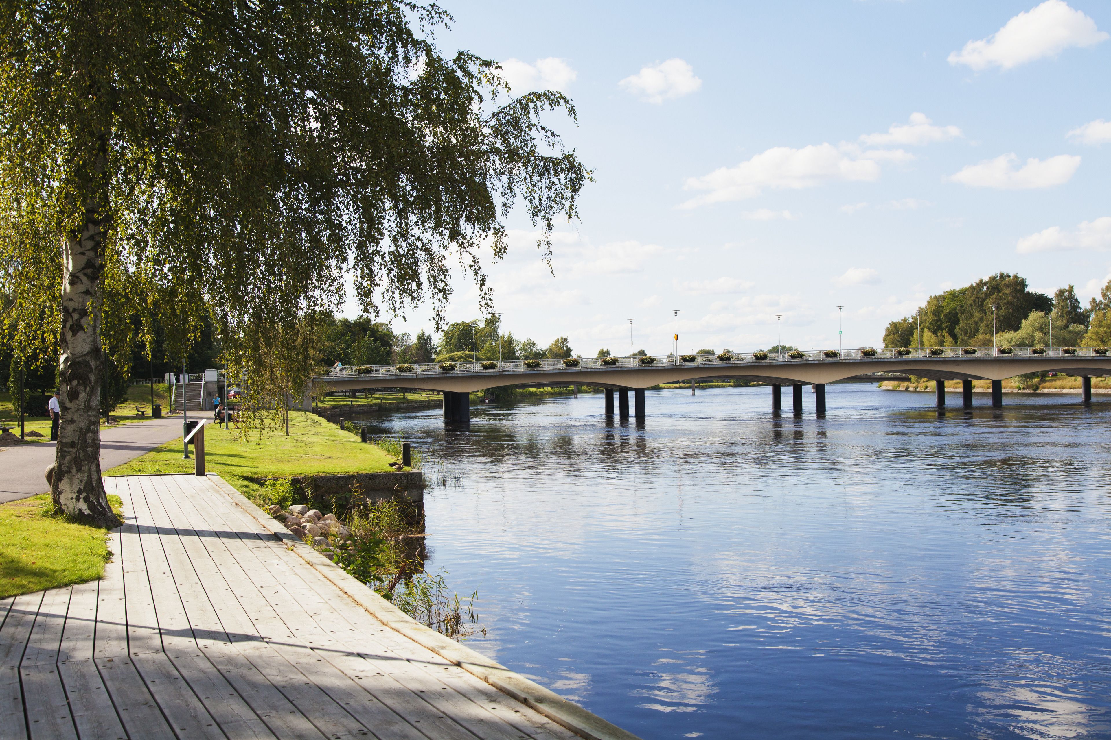 A wooden boardwalk along a river with a bridge in the background.
