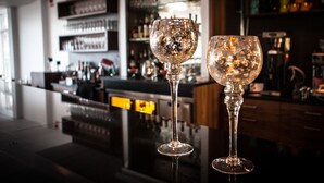 Dos copas de cristal transparente sobre una barra reflectante, con un fondo borroso de estantes y botellas.