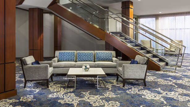 A hotel lobby with a staircase, seating area, and patterned carpet. - Courtyard by Marriott Woburn/Boston North (Woburn)