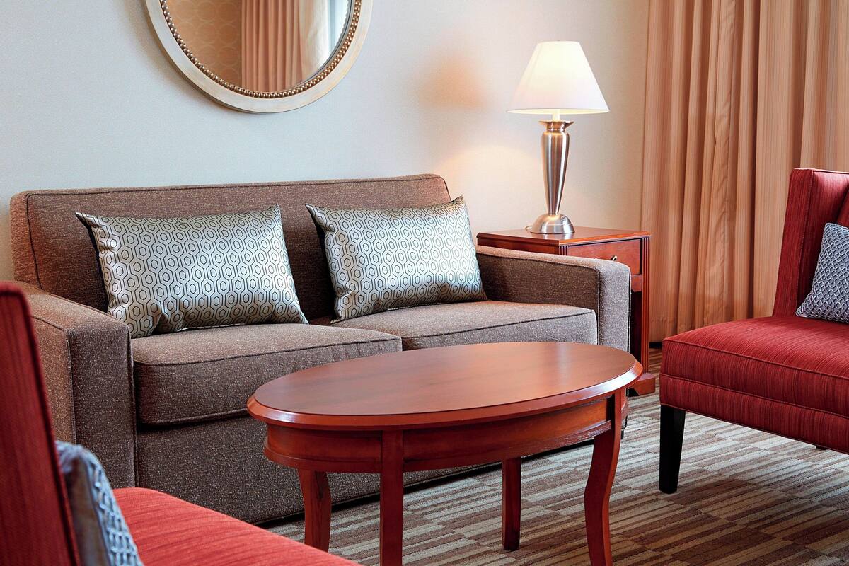 A hotel room with a sofa set, a round wooden coffee table, a lamp, and a mirror on the wall.