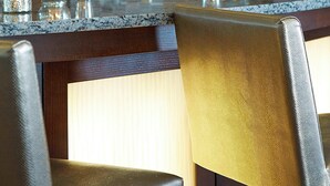 A bar area with a tiled backsplash, a glass shelf displaying various bottles, and a seating area with chairs and a table. - Courtyard by Marriott Woburn/Boston North (Woburn)