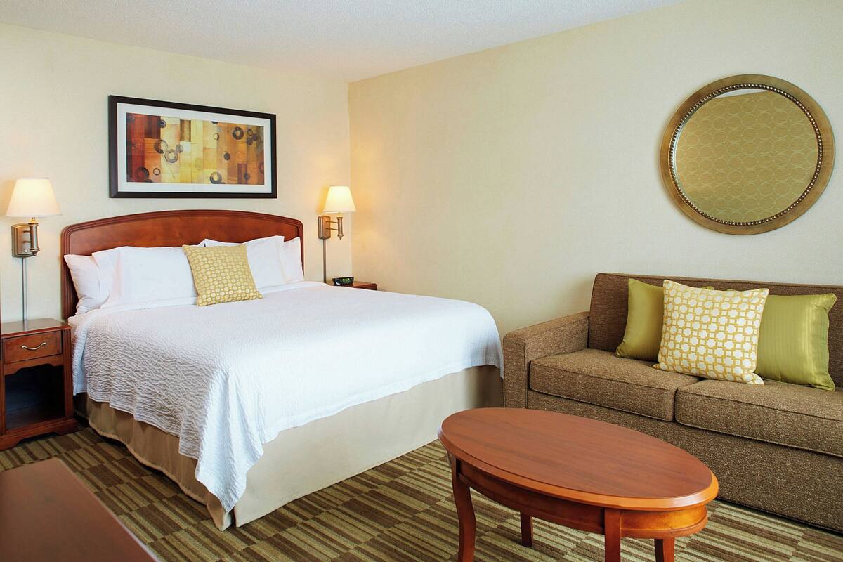 A hotel room with a bed, a sofa, a round mirror, and two wall-mounted lamps.
