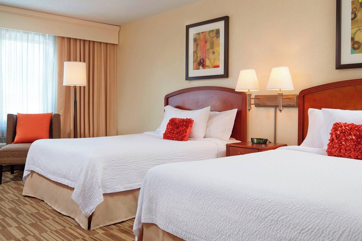 A hotel room with two beds, each with white bedding and red decorative pillows. There are two bedside tables with lamps, a window with curtains, and a framed picture on the wall.