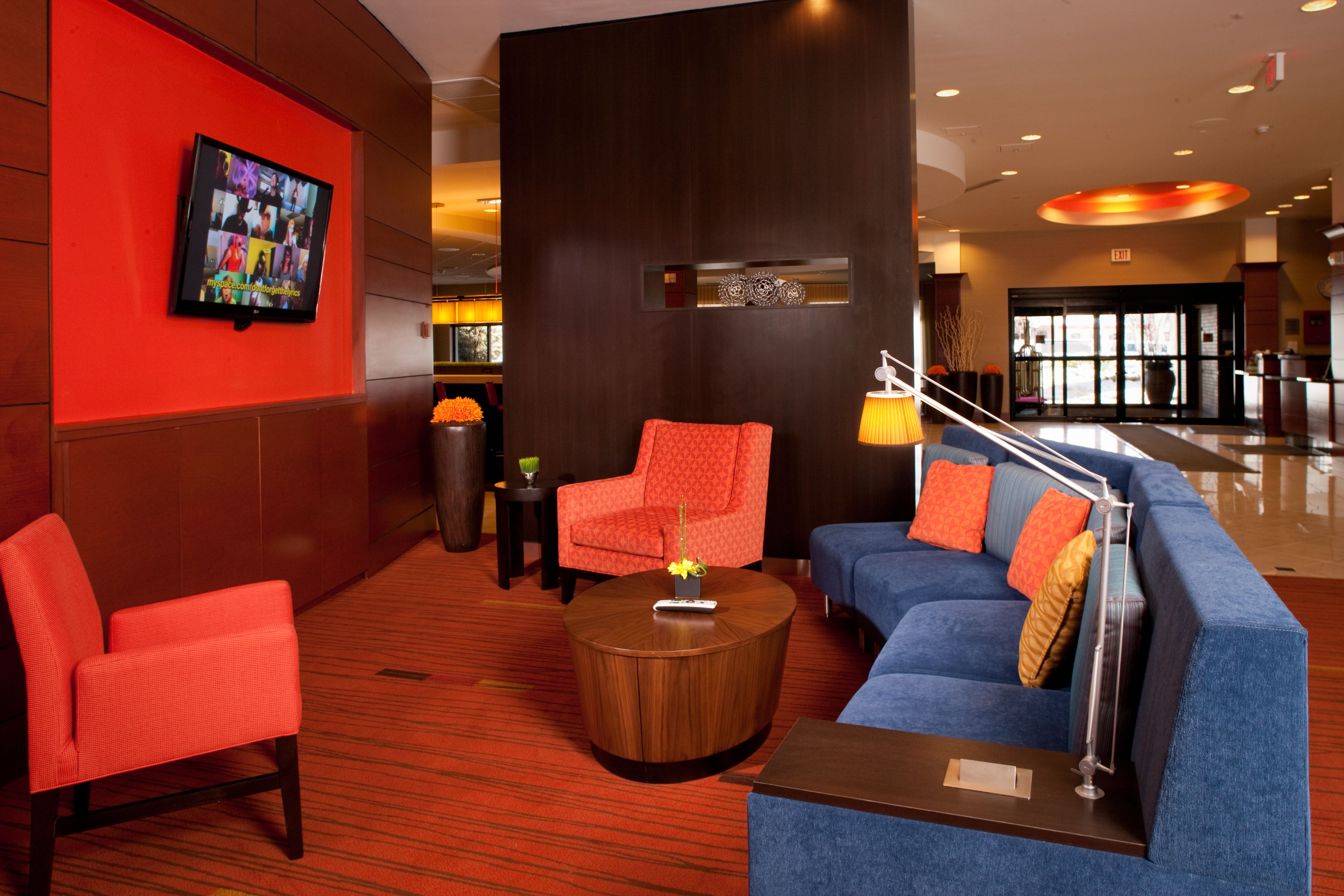 A lobby with a red wall, a television mounted on it, a red armchair, a blue sofa with orange pillows, and a round wooden coffee table.