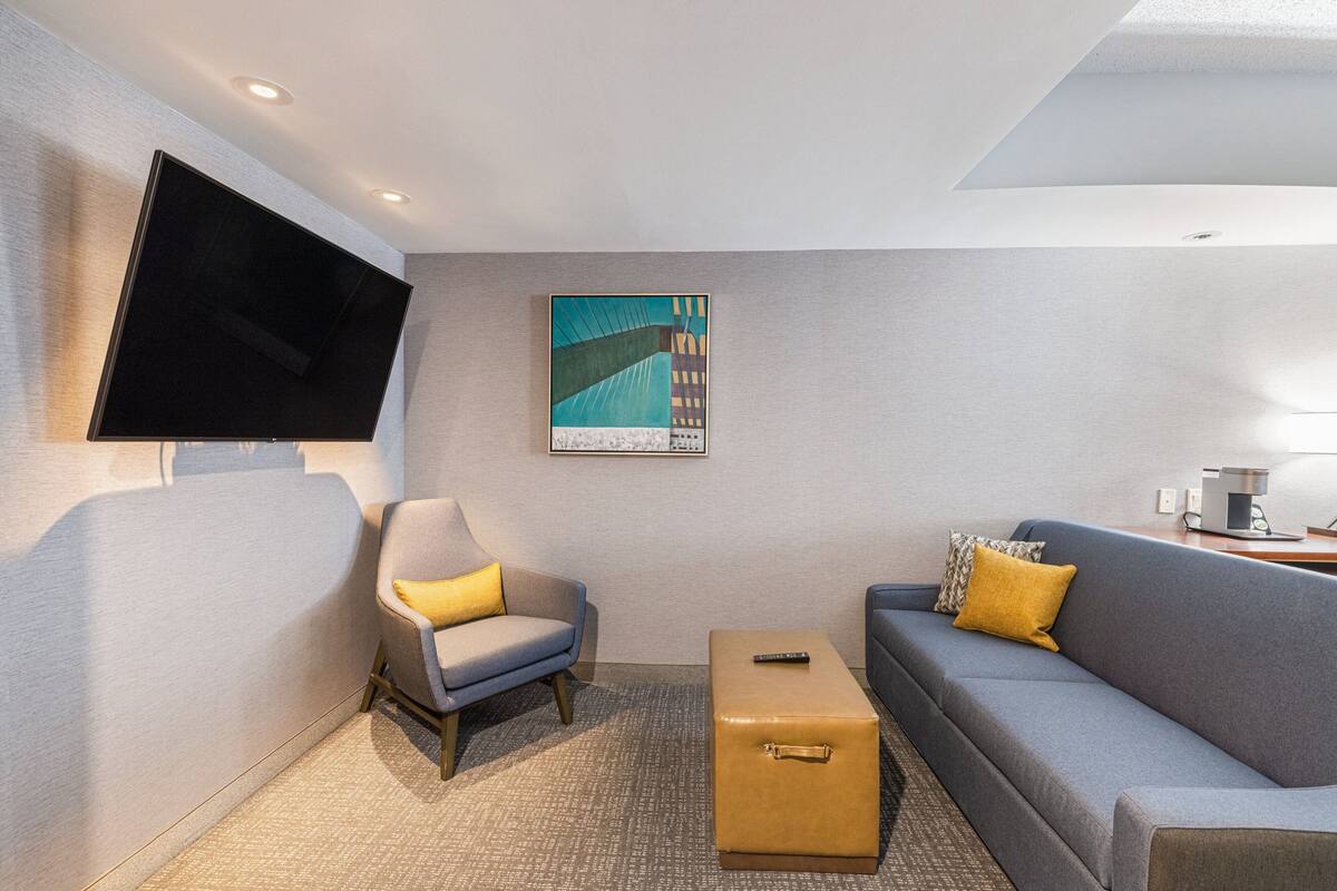 A modern hotel room with a flat-screen TV mounted on the wall, a sofa with yellow cushions, a small ottoman, and a painting above the sofa.