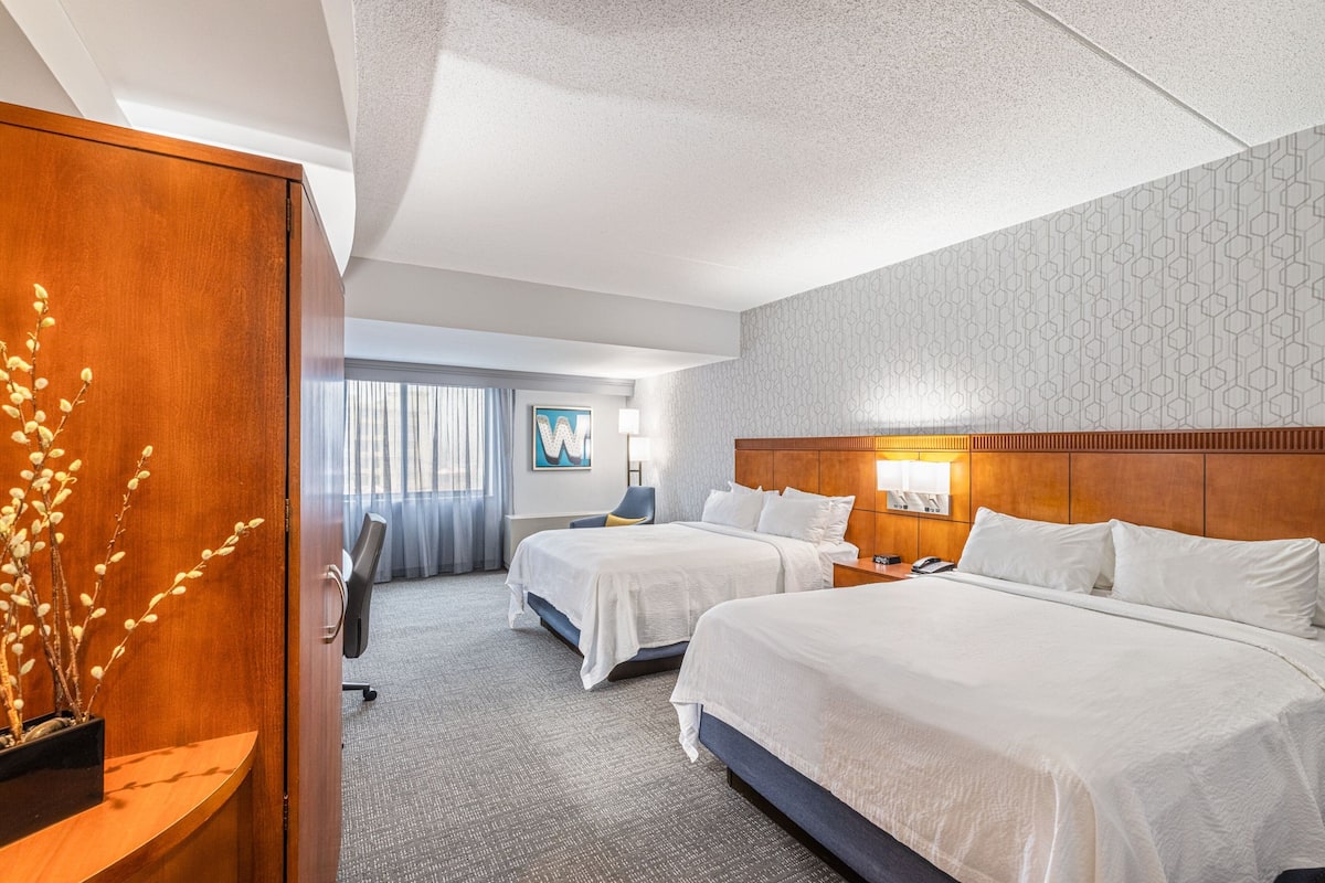 A hotel room with two beds, a wooden headboard, a desk with a lamp, and a view of the hallway.