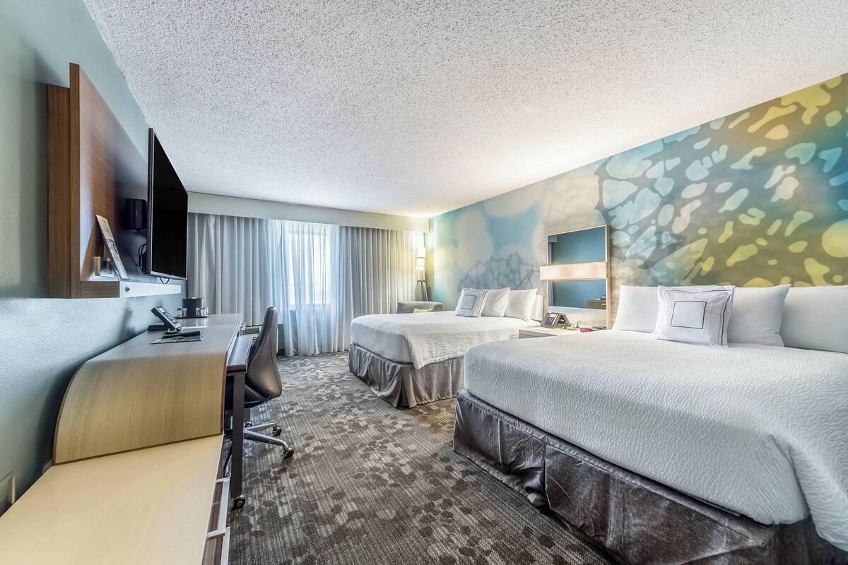 Une chambre d’hôtel équipée d’un bureau, de deux lits, d’une télévision et d’une grande fenêtre avec des rideaux.