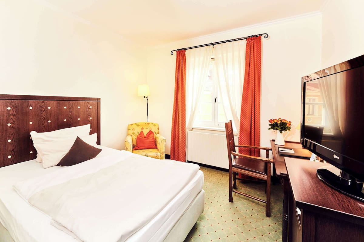 A hotel room with a bed, a desk with a television, a chair, and a window with curtains.