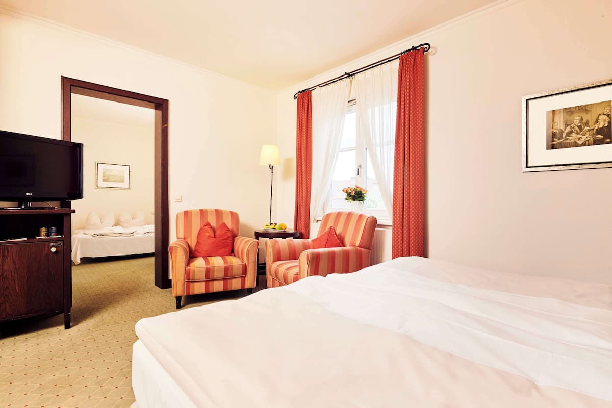 A hotel room with a bed, two striped armchairs, a desk with a lamp, a television, and a framed picture on the wall.