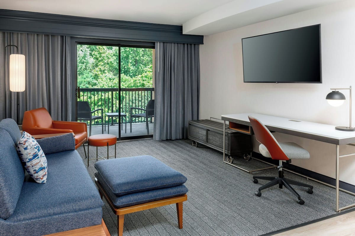 A modern hotel room with a sofa, a desk, a TV, and a balcony with outdoor furniture.