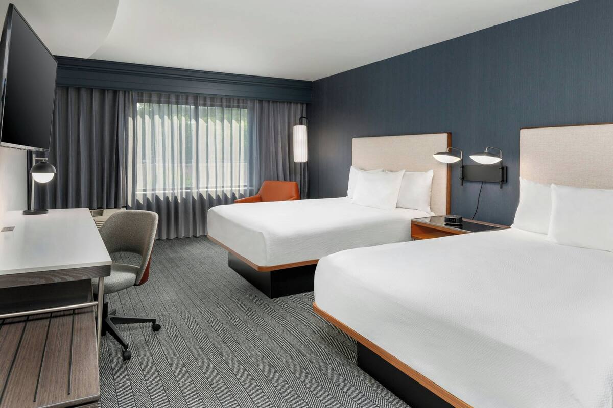 A hotel room with two beds, a desk, a chair, and a TV.