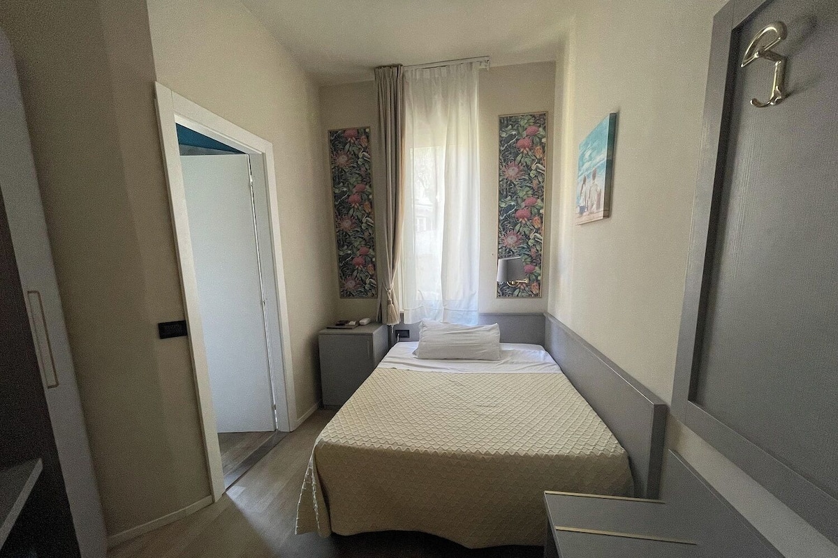 A narrow hotel room with a bed, a small bedside table, a nightstand, and a door.