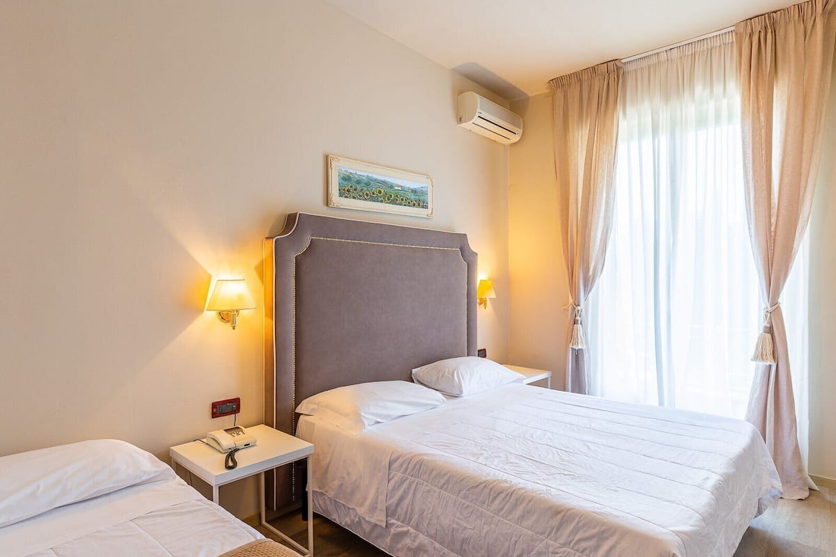 A hotel room with a large bed, a smaller bed, a bedside table with a phone, a wall-mounted air conditioner, and a window with curtains.