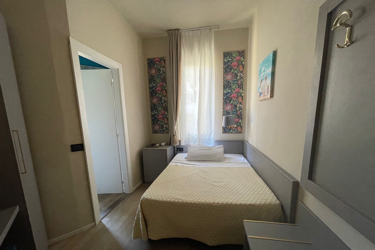 A hotel room with a bed, a desk, a chair, a nightstand, and a window with curtains.