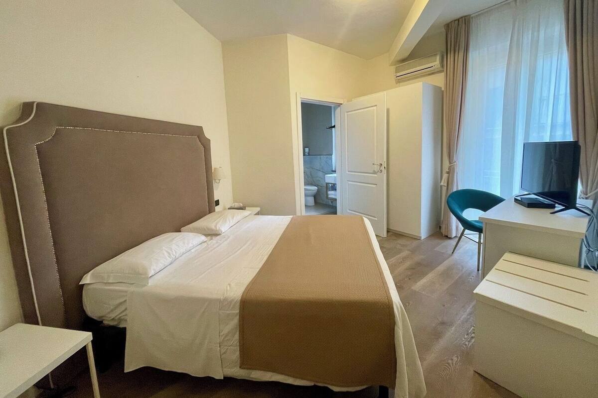 A hotel room with a bed, a desk, a chair, a television, and an air conditioning unit.