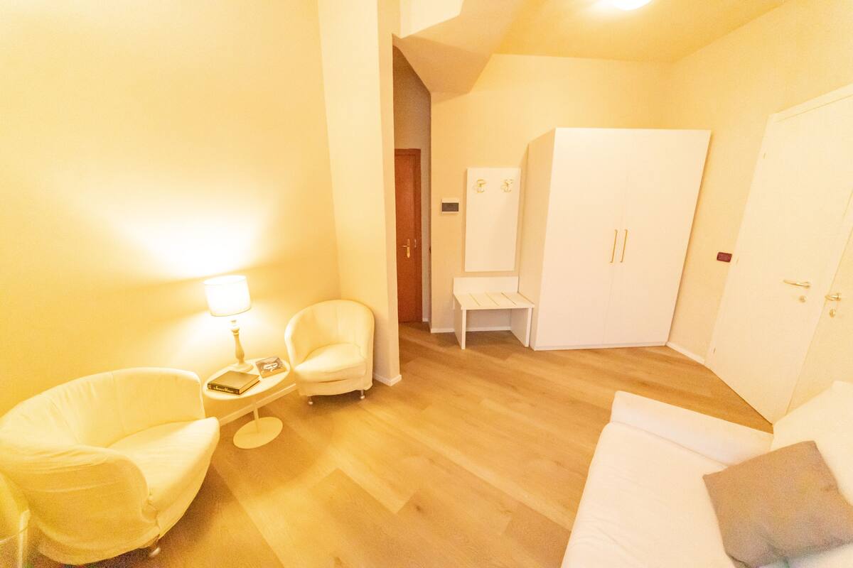 A hotel room with a wooden floor, a white sofa, a white armchair, a small table with a lamp, and a wardrobe.