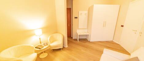 Una habitación de hotel con piso de madera, un sofá blanco, un sillón blanco, una mesita con lámpara y un armario.
