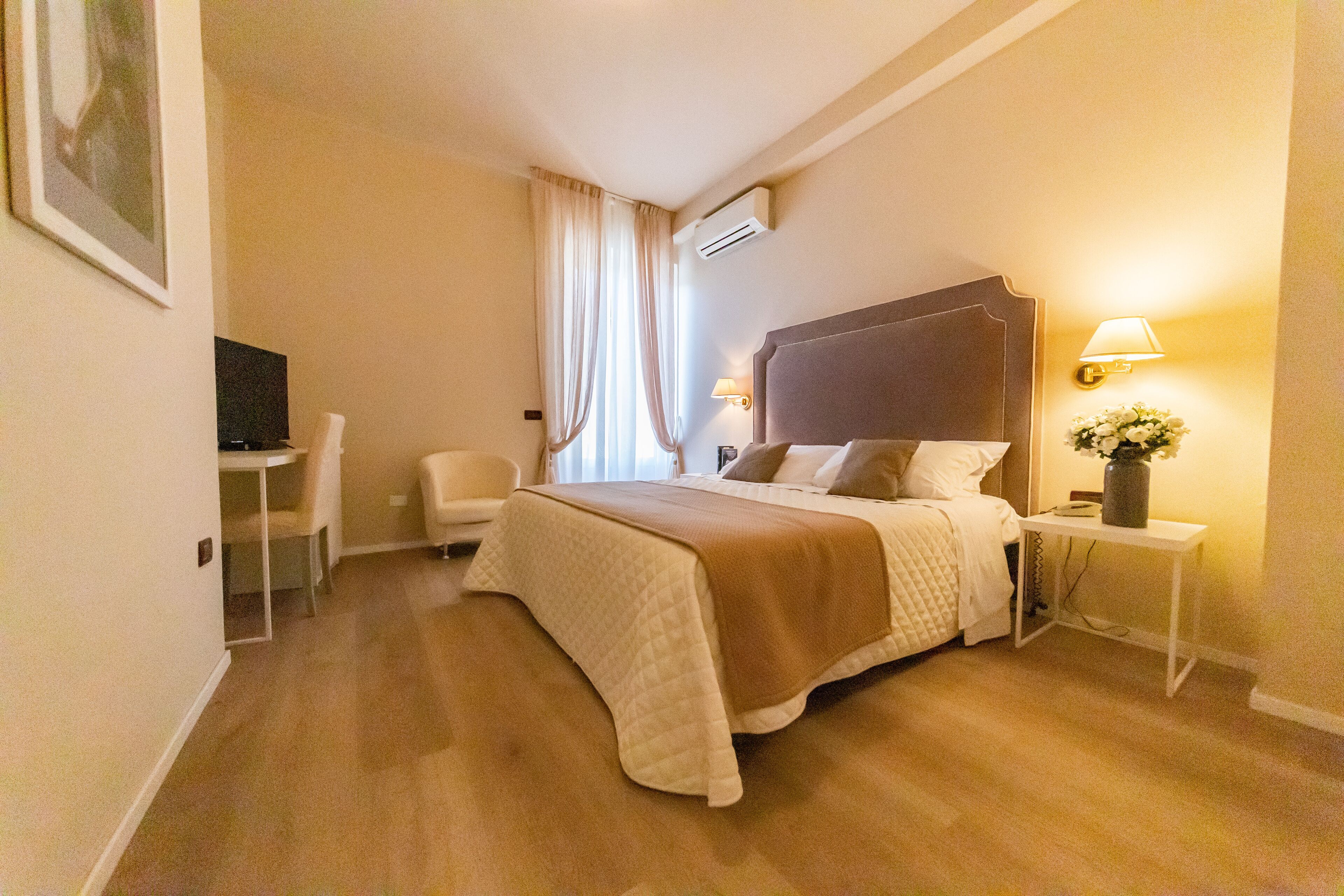 A hotel room with a bed, a desk, a chair, a TV, a lamp, and a vase with flowers.