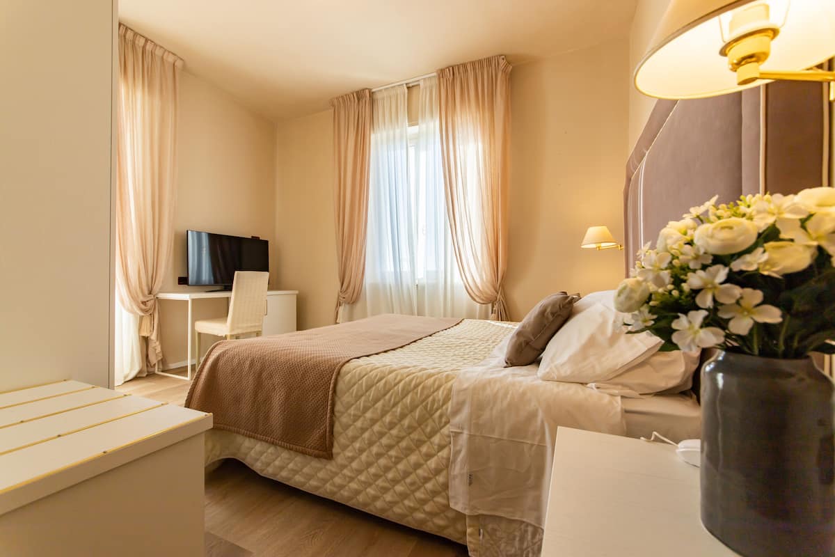 A hotel room with a bed, bedside table, chair, TV, and a vase with flowers.