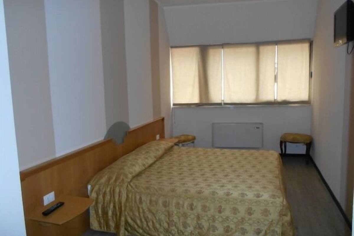 A small, simple room with a bed, a nightstand, a chair, and a TV mounted on the wall.