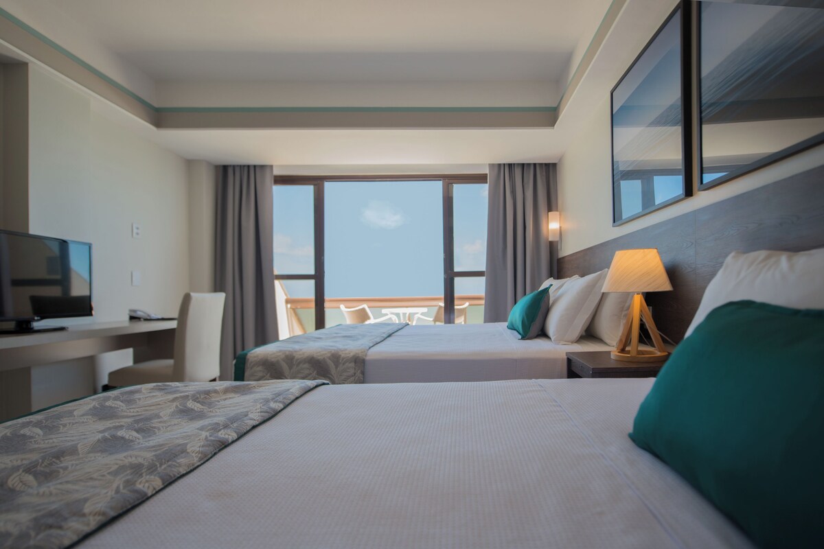 A hotel room with two beds, a desk, a television, and a balcony with a view.