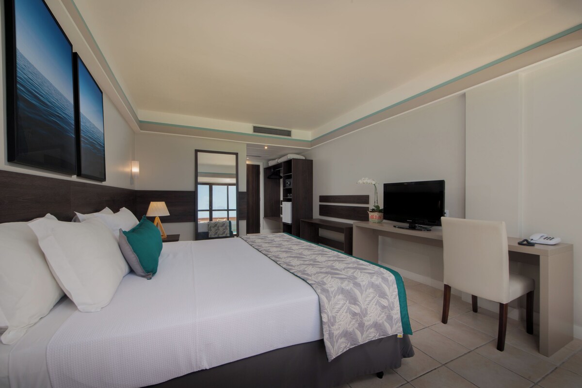 A hotel room with a large bed, a desk with a television, and a view of the sea through the window.